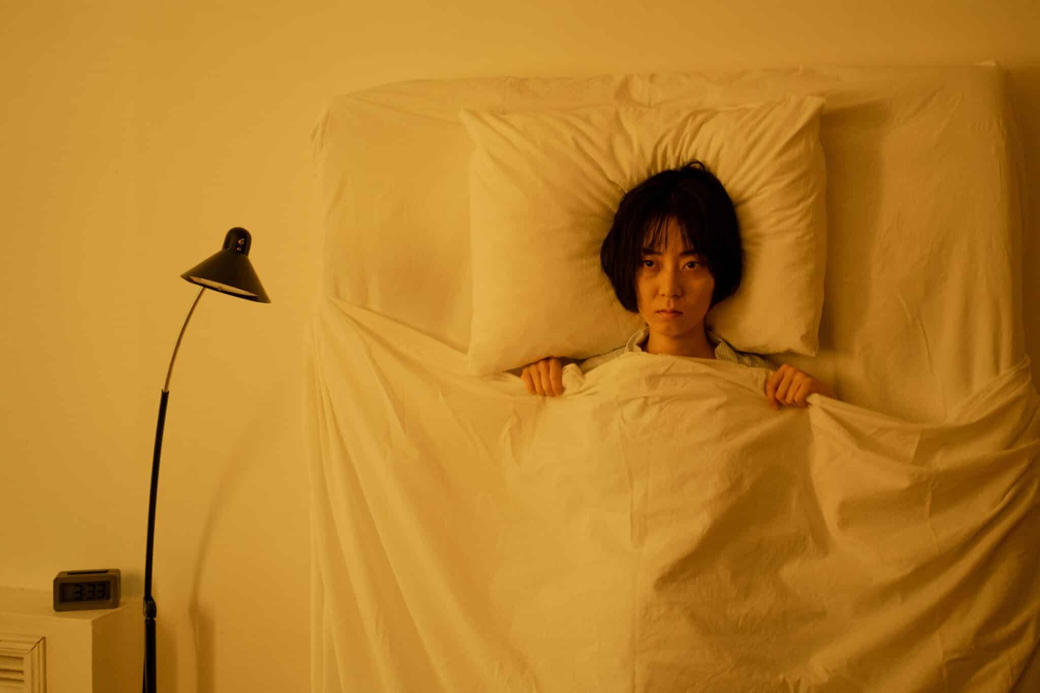 Woman lying under a blanket with a moody expression in a dimly-lit bedroom.